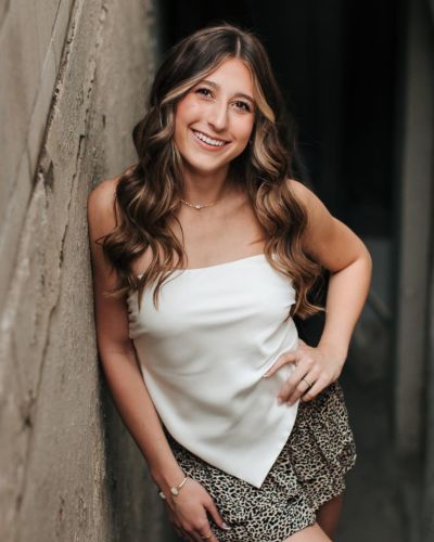 A young woman with long wavy brown hair smiles while leaning against a textured wall. She is wearing a white strapless top and a patterned skirt, with one hand on her hip and the other touching the wall.