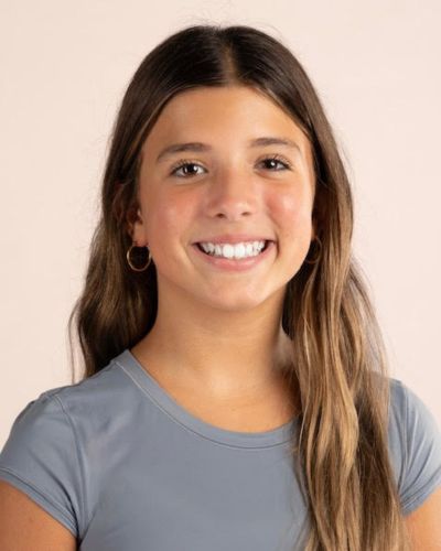A young girl with long brown hair, wearing a gray short-sleeve top and gold hoop earrings, smiles in front of a plain light background.