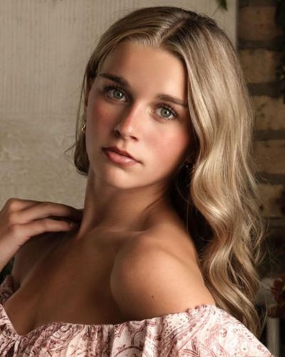 A young woman with long, wavy blonde hair poses indoors. She wears an off-the-shoulder, patterned top and gazes softly at the camera, her hand resting near her neck against a neutral, softly lit background.