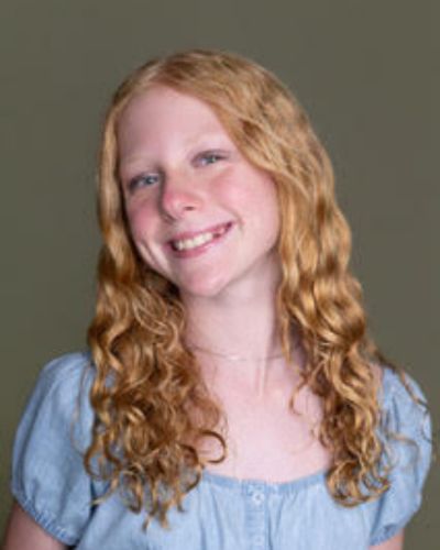 A young person with long, wavy red hair smiles at the camera. They are wearing a light blue, short-sleeved, button-up shirt, and are standing in front of a plain, olive-green background.