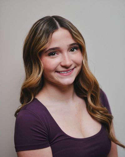 A young woman with long, wavy brown hair wearing a purple top smiles at the camera against a plain, light-colored background.