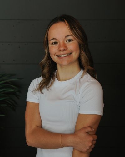 A young woman with wavy brown hair stands in front of a dark wall, smiling, and wearing a short-sleeve white shirt. She has one arm crossed and her other hand resting on her arm. A green plant is partially visible beside her.