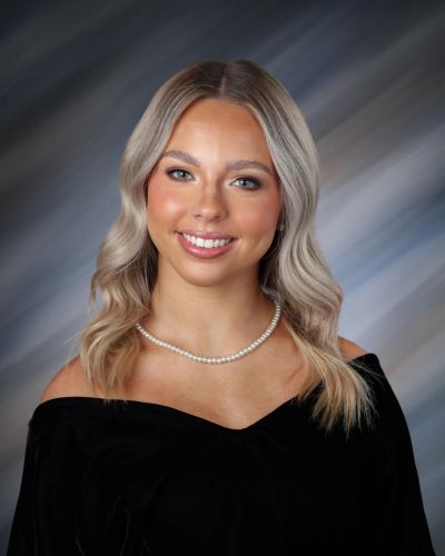 A young woman with wavy blonde hair, wearing a black off-the-shoulder top and a pearl necklace, smiles in front of a softly blurred, multicolored background.