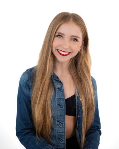 A young woman with long blonde hair smiles brightly, wearing a blue denim jacket over a black top, against a plain white background.