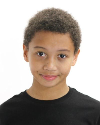 A young child with short curly hair, wearing a black shirt, is smiling softly and looking directly at the camera against a plain white background.