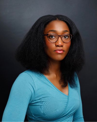A woman with long, natural black hair and glasses wears a teal wrap top and looks at the camera with a neutral expression against a dark background.