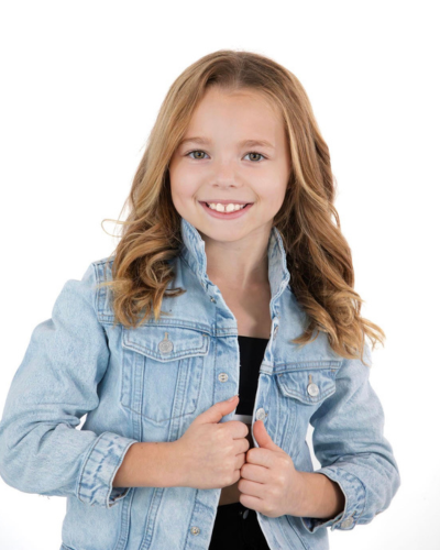 A young girl with long, wavy blonde hair smiles at the camera. She is wearing a light blue denim jacket over a black shirt, standing against a plain white background.