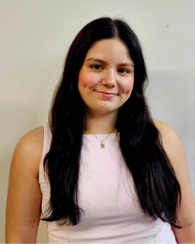 A young woman with long dark hair wearing a pale pink sleeveless top smiles gently in front of a plain light-colored background.