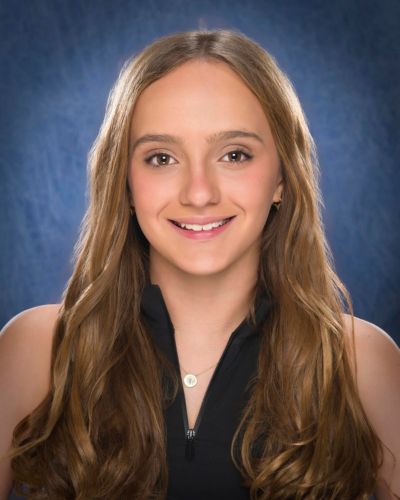 A young person with long, wavy light brown hair smiles at the camera. They are wearing a black sleeveless top and a necklace with a round pendant, set against a blue textured background.