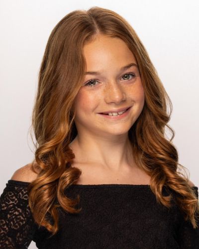 A young girl with long, wavy light brown hair smiles at the camera. She is wearing an off-the-shoulder black lace top and is posed against a plain, light background.