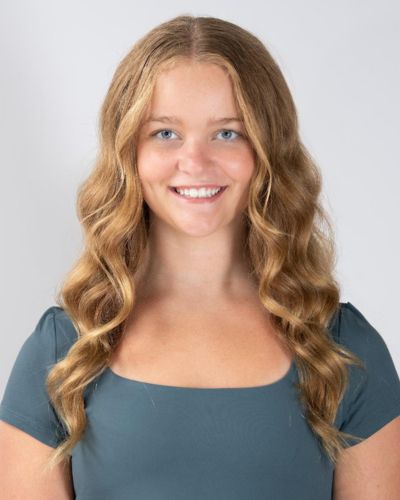 A young woman with long, wavy blonde hair and blue eyes smiles at the camera. She is wearing a short-sleeved, square-necked, teal top against a plain light background.