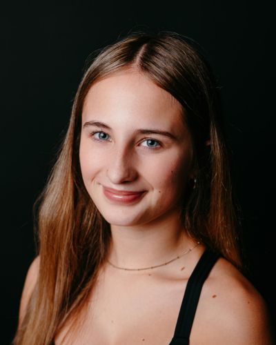 A young woman with long light brown hair and blue eyes smiles gently at the camera. She is wearing a black top and a delicate necklace. The background is solid black.