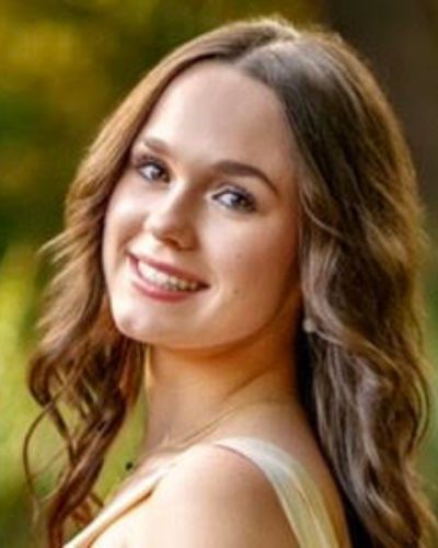 A young woman with long, wavy brown hair smiles at the camera. She is outdoors, wearing a light-colored top, with a blurred green background suggesting a natural setting.