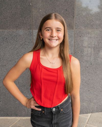 A smiling young girl with long brown hair wears a sleeveless red top and black pants, standing with one hand on her hip against a gray stone wall background.