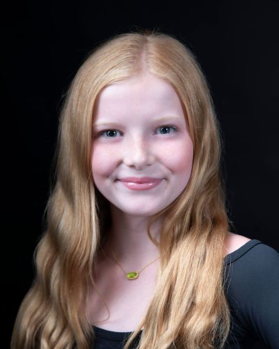 A young girl with long, wavy blonde hair and fair skin smiles softly. She is wearing a black top and a gold necklace with a small green pendant, set against a dark background.