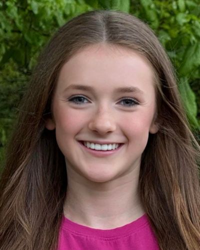 A teenage girl with long brown hair smiles at the camera. She is wearing a pink shirt and standing outdoors with green foliage in the background.