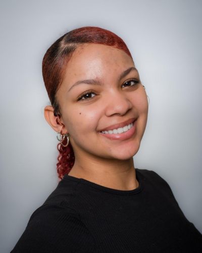 A young woman with reddish-brown hair tied back smiles at the camera. She is wearing a black top and gold hoop earrings, standing against a plain light gray background.