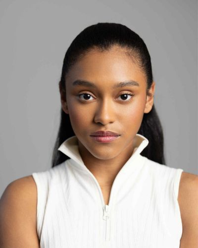 A young woman with medium skin tone and straight, dark hair pulled back, wearing a sleeveless white top with a zipped collar, looks directly at the camera against a plain gray background.