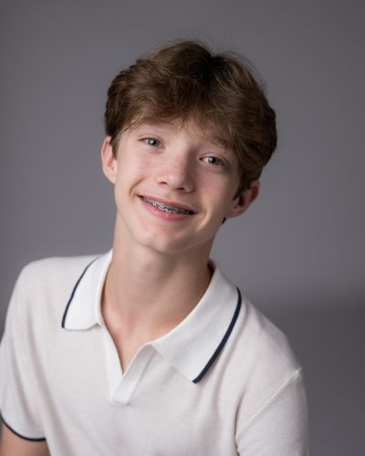 A smiling teenage boy with light brown hair and braces, wearing a white collared shirt with dark trim, poses in front of a plain gray background.