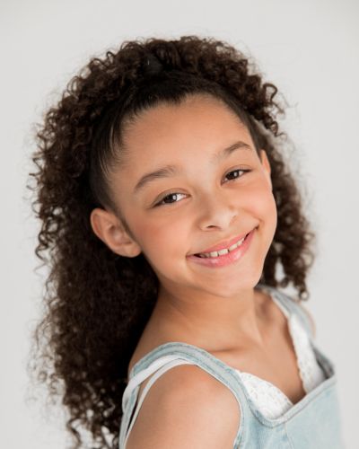 A young girl with curly brown hair smiles warmly at the camera. She is wearing a light blue top with white straps, and the background is plain and light-colored.