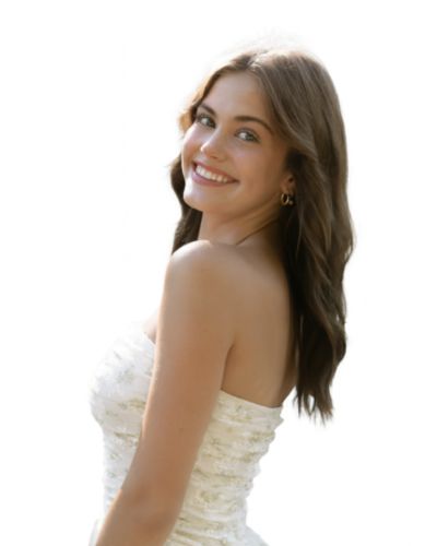 A young woman with long brown hair smiles at the camera, wearing a strapless white dress and hoop earrings, standing against a plain white background.