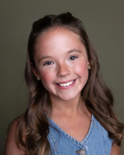 A young girl with long brown hair styled in loose curls smiles at the camera. She is wearing a sleeveless denim top and gold earrings, set against a plain olive-green background.