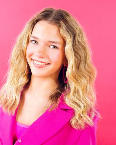 A young woman with wavy blonde hair smiling at the camera, wearing a bright pink blazer and hoop earrings, standing in front of a solid pink background.