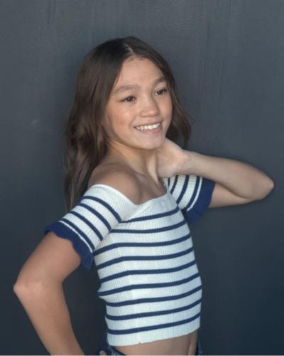 A young girl with long brown hair smiles and poses with one hand behind her neck. She wears an off-shoulder, short-sleeve, white and navy striped top, standing in front of a plain dark gray background.