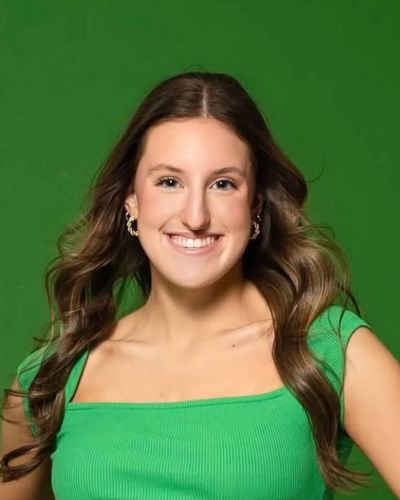 A young woman with long, wavy brown hair smiles at the camera. She is wearing a green, short-sleeved top and is posed in front of a solid green background.
