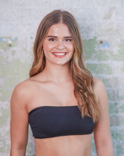 A young woman with long, wavy brown hair is smiling at the camera. She is wearing a black bandeau top and standing in front of a textured, light-colored wall with some green and white patches.