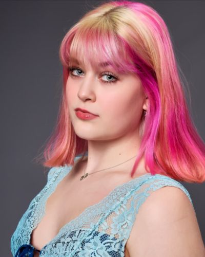 A young woman with straight, shoulder-length pink and blonde hair poses against a gray background. She wears a light blue lace top and a silver necklace, looking slightly to the side with a neutral expression.