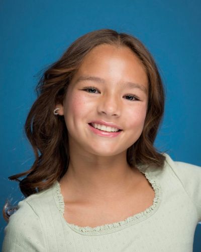 A young girl with long brown hair and light skin smiles in front of a solid blue background, wearing a light green top with a ruffled neckline.