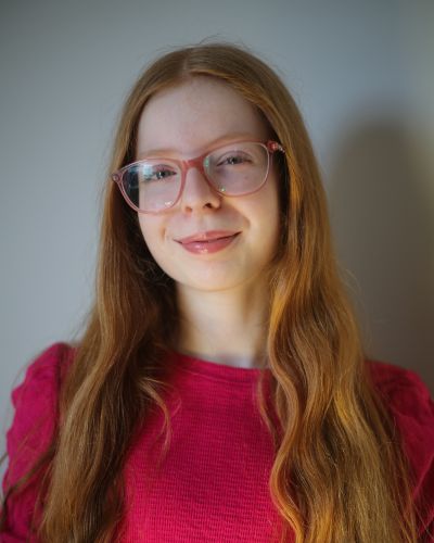 A young person with long, wavy red hair and pink glasses is smiling softly at the camera. They are wearing a bright pink top and standing against a neutral background.
