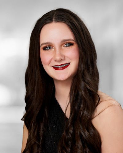 A young woman with long, wavy dark hair smiles at the camera. She is wearing dark red lipstick, eye makeup, and a black, sleeveless top. The background is blurred and gray.