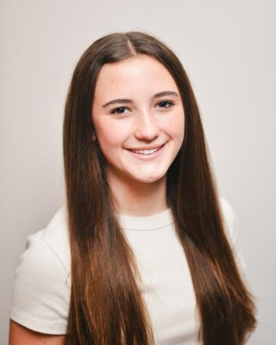 A young woman with long, straight brown hair and a light-colored top smiles at the camera against a plain, light background.