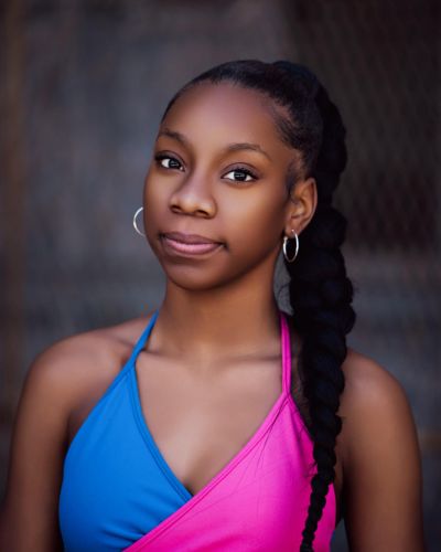 A young girl with hoop earrings and a long braided ponytail, wearing a sleeveless blue and pink top, stands against a blurred outdoor background, looking calmly at the camera.