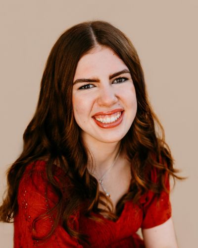 A young woman with long brown hair and a bright smile wears a red top, sitting against a plain beige background.