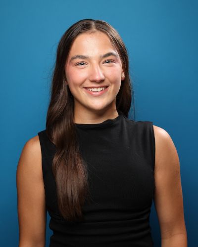 A young woman with long brown hair wearing a sleeveless black top smiles at the camera in front of a solid blue background.