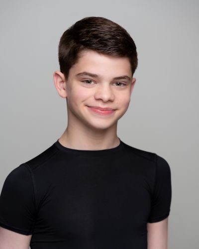 A young boy with short dark hair smiles at the camera. He is wearing a fitted black t-shirt and standing against a plain light gray background.