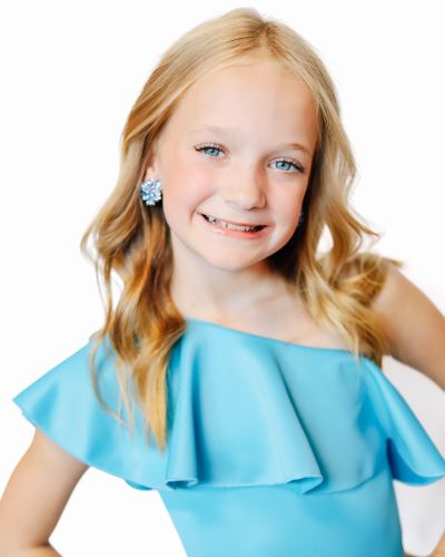 A young girl with long blonde hair, wearing a light blue off-the-shoulder dress and blue flower-shaped earrings, smiles at the camera against a plain white background.