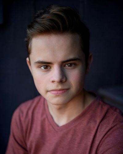 A young man with short brown hair and fair skin wears a mauve V-neck shirt, looking directly at the camera with a neutral expression. The background is dark and out of focus.