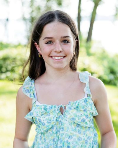 A young girl with long brown hair, wearing a light blue floral sleeveless dress, smiles outdoors with green foliage and trees blurred in the background.