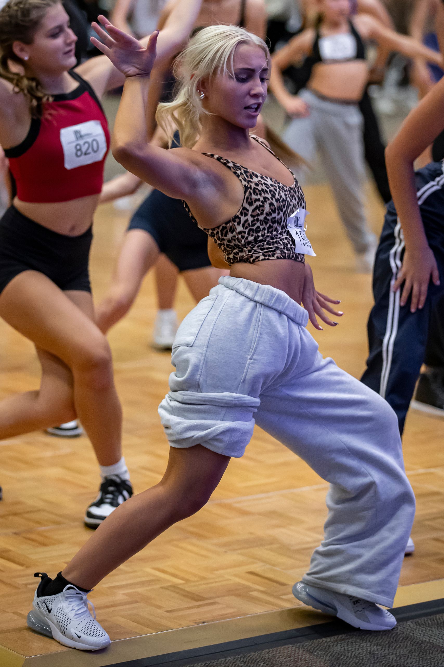 A young woman in a leopard-print crop top and baggy sweatpants dances energetically in a group audition, striking a dynamic pose with one arm raised and bent, surrounded by other dancers on a wooden floor.