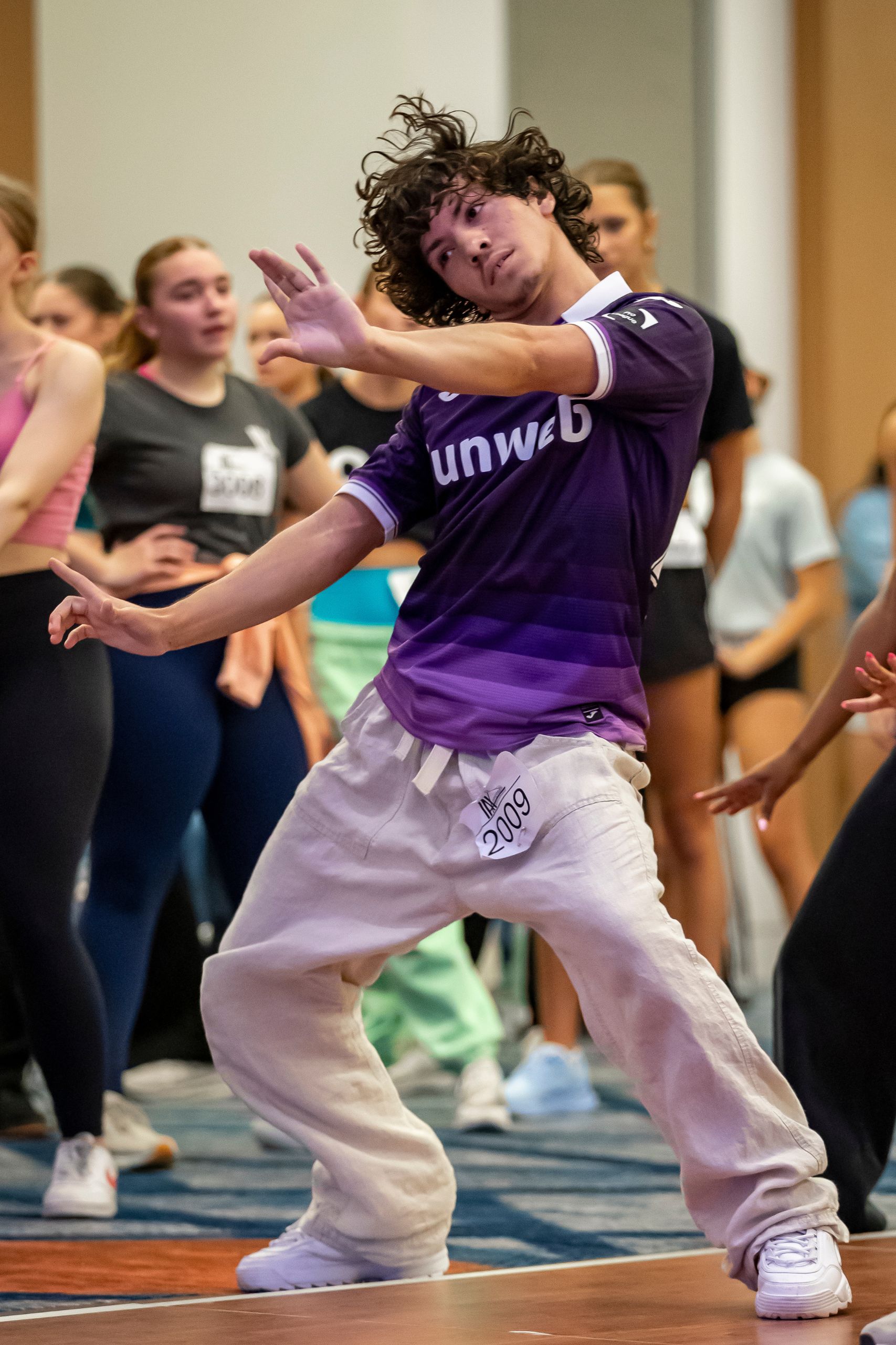 A young person with curly hair, wearing a purple jersey and white pants, dances energetically in a group, striking a dramatic pose with arms raised and bent, while others watch in the background.