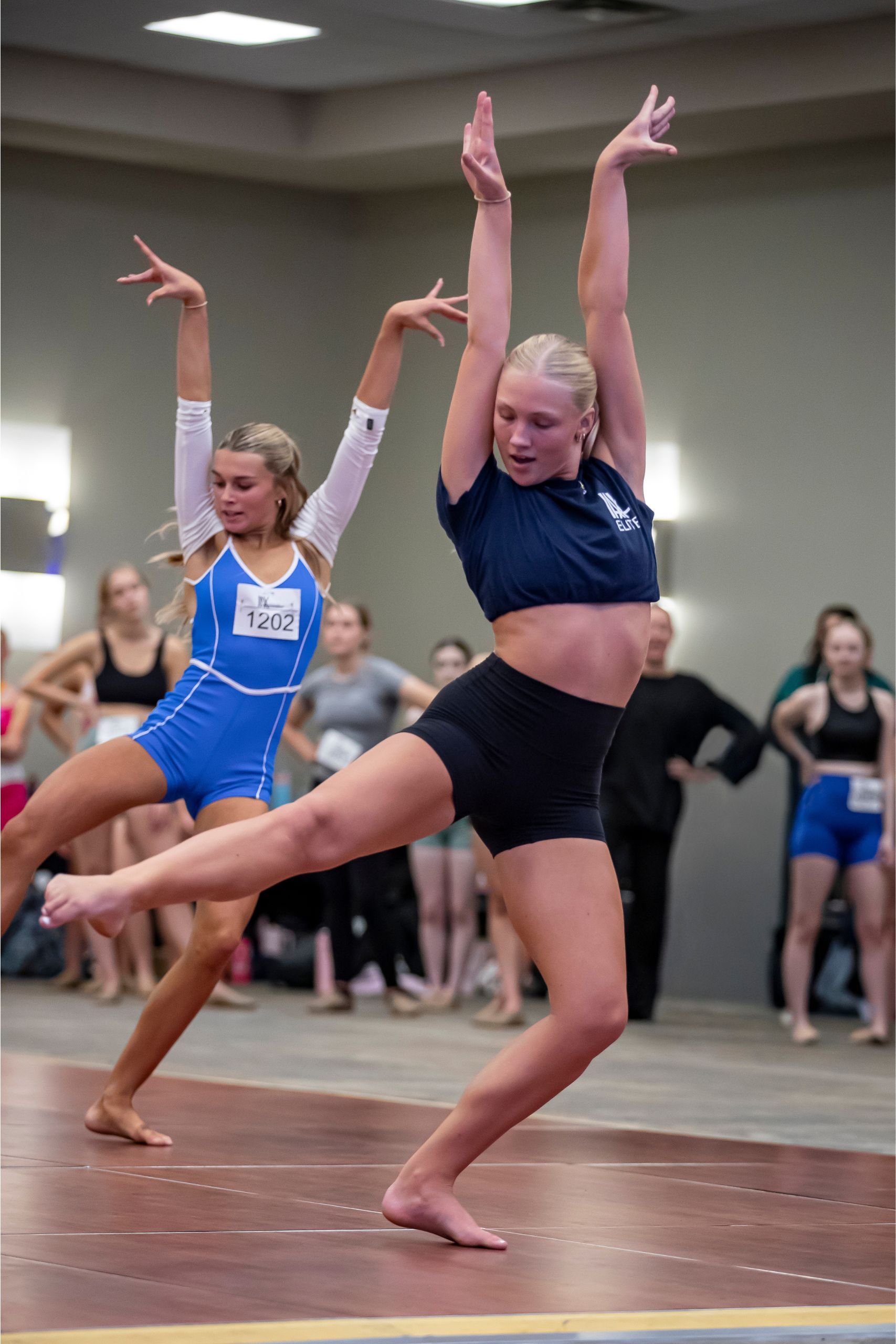 Two young female dancers perform a contemporary routine indoors, wearing dance attire and striking expressive poses with arms raised. Other dancers and onlookers watch in the background.