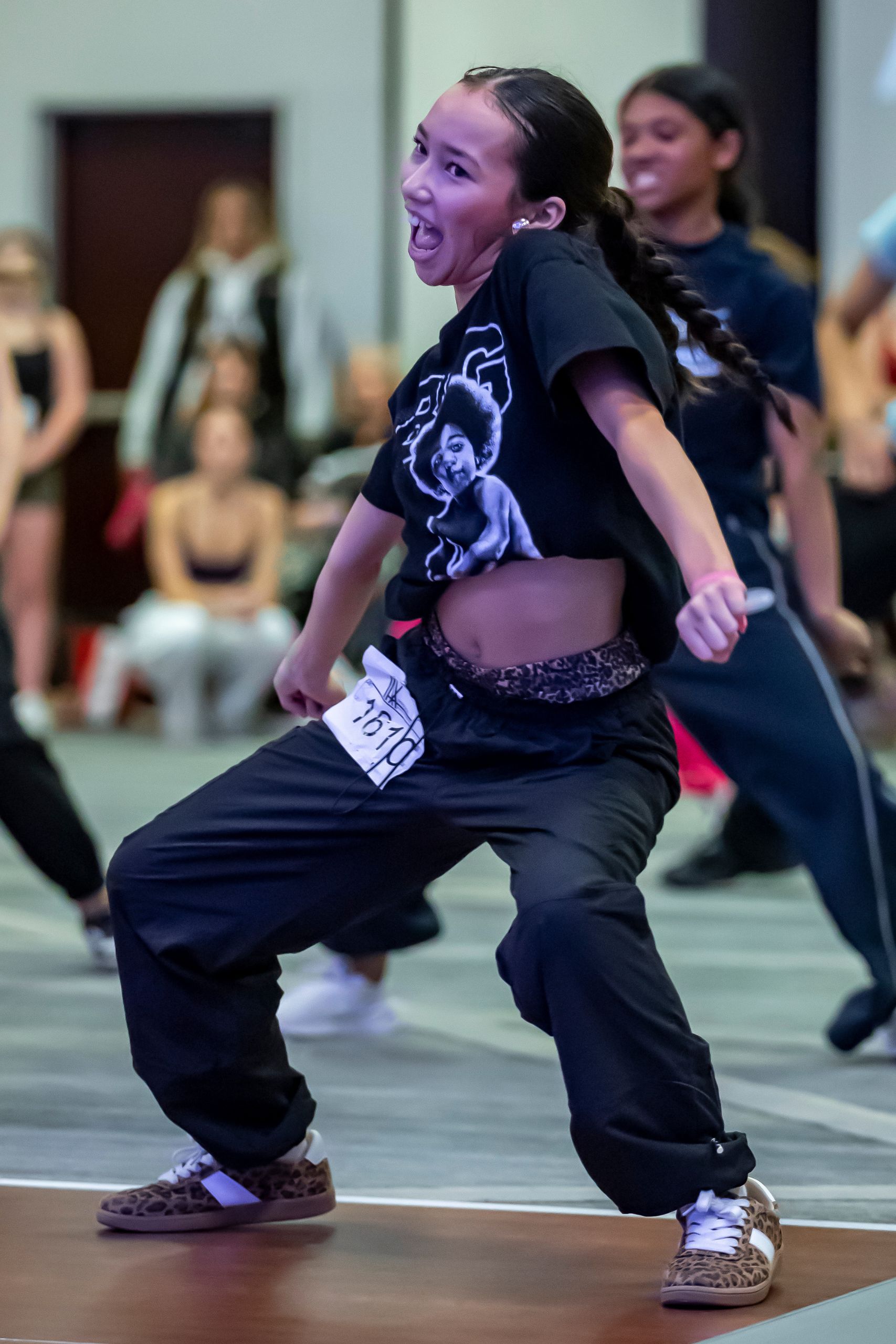 A young woman in loose black clothing and sneakers energetically dances at an indoor event. She wears a number tag on her pants, and people in the background watch her performance.