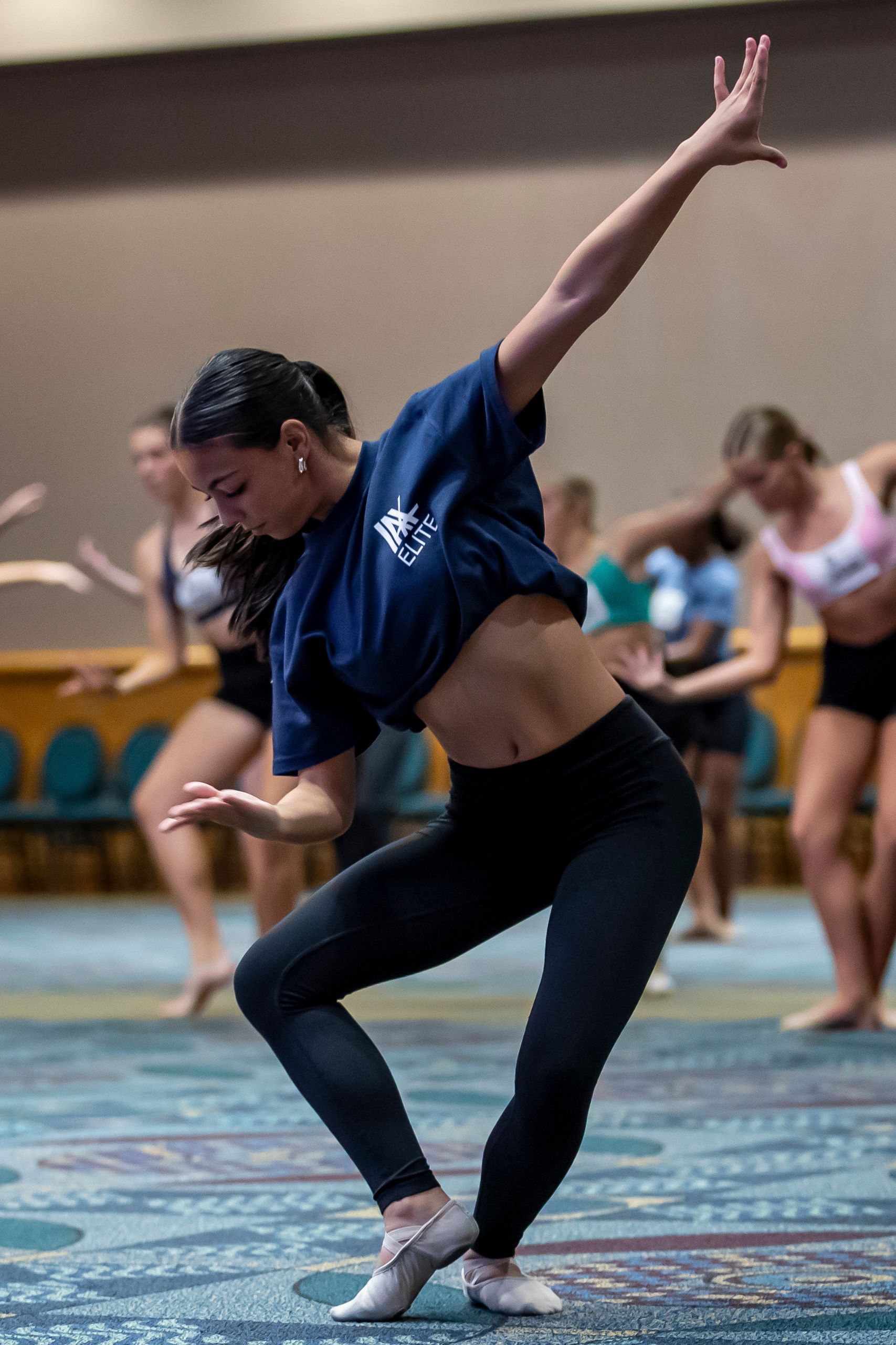 A young woman in a navy crop top and black leggings performs a contemporary dance move in a studio, while several other dancers practice in the background.