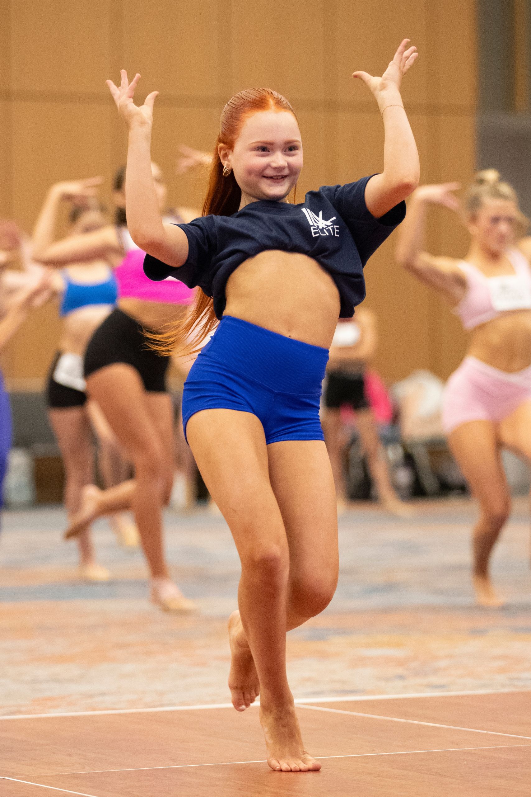 A young girl with red hair, wearing a navy t-shirt and blue shorts, dances energetically indoors. Other dancers in colorful outfits perform in the background on a gymnasium floor.