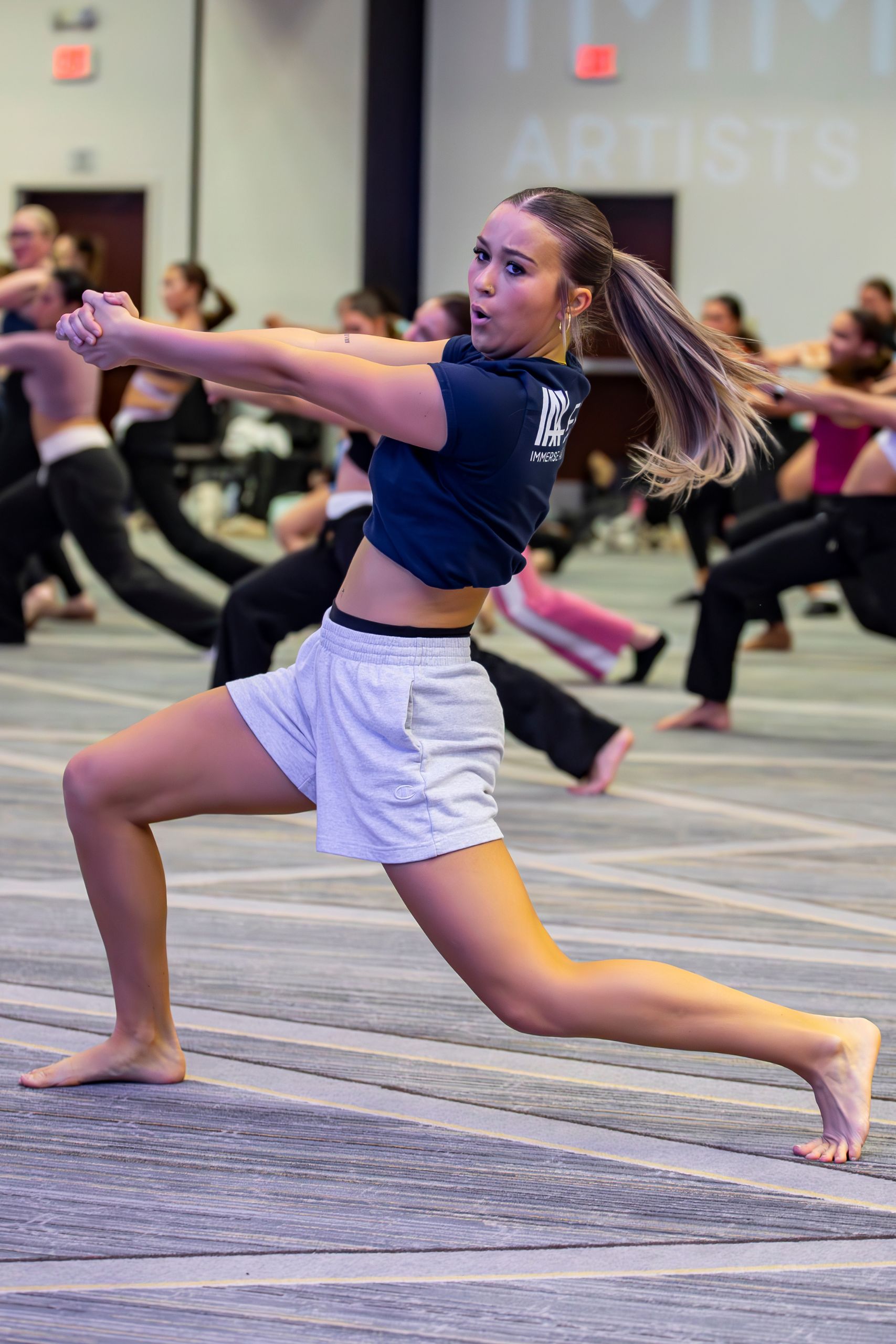 A young woman in athletic wear performs a dynamic dance move in a large room with other dancers in the background, all practicing together. She stretches her arms and legs, looking focused and energetic.