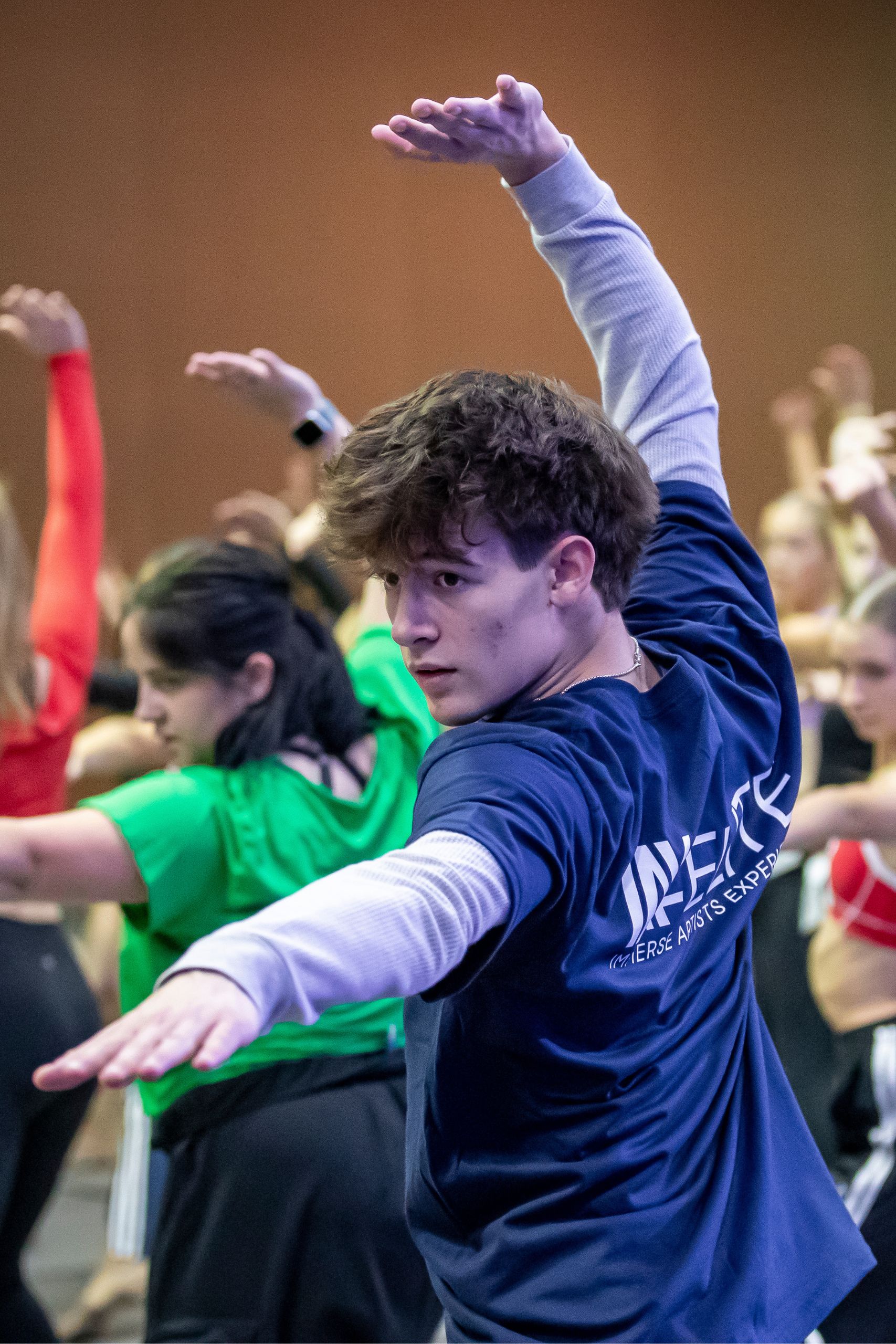 A young person in a navy t-shirt and long sleeves extends their arms in a dance pose, surrounded by a group of dancers practicing similar movements in a studio.